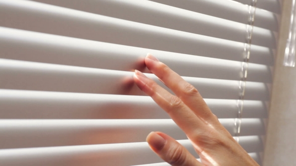 Female Hand Separating Slats Of Venetian Blinds With a Finger To See Through