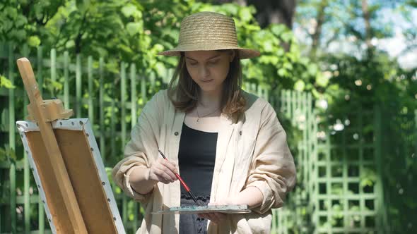 Young Woman Artist in Brown Hat Stands at Easel and Paints