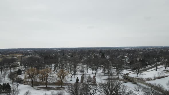 Aerial view over park with creek winding through, bridge and snow on the ground.