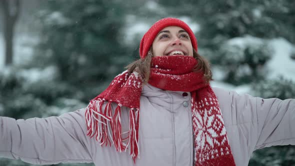 Beautiful Smiling Woman in Red Beanie and Scarf Standing in Forest at Snowy Winter Day Outdoors