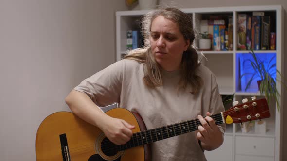 A woman with a serious face plays an acoustic guitar sitting in a room