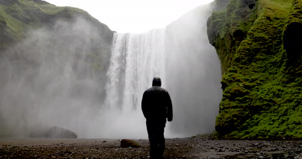 Skogafoss waterfalls in Iceland with man in rain jacket walking towards falls in slow motion.
