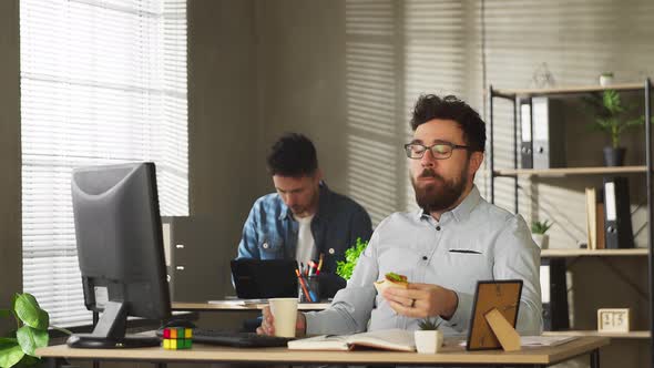 Young Man in Glasses and Casual Outfit Eating Sandwich While Working in Office