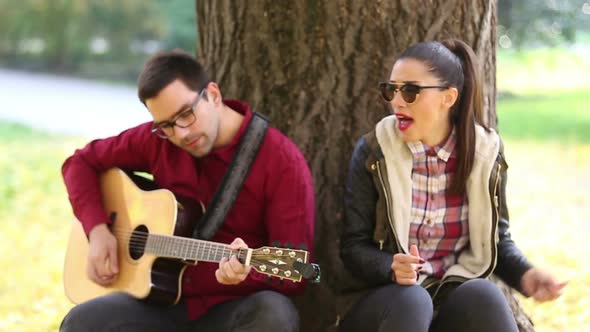 Woman Singing And Man Playing Guitar While Sitting On A Tree In Park 2