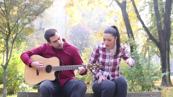 Man Playing Guitar And Singing With Women While Sitting On Bench In Park 1