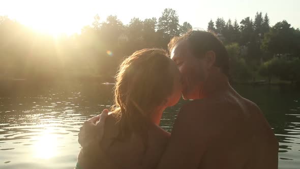 Couple In Love Kissing And Smiling While Sitting By The River At Sunset