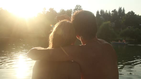 Couple In Love Taking Selfies And Kissing While Sitting By The River At Sunset