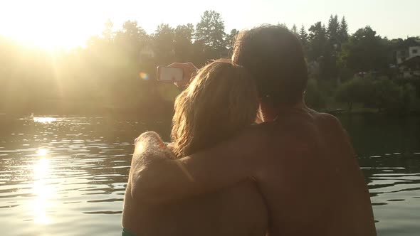 Couple In Love Taking Selfies And Laughing While Sitting By The River At Sunset