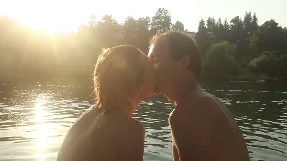 Couple Looking At Each Other And Kissing While Sitting By The River At Sunset