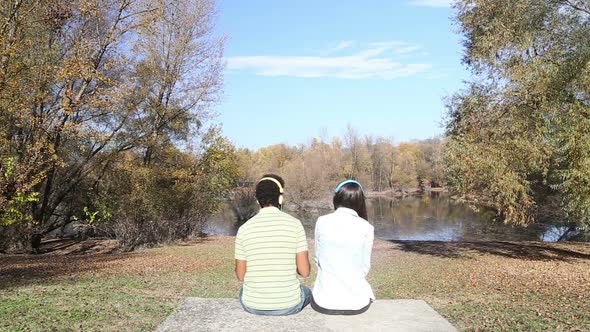 Man And Woman Listening To Music Using Headphones While Sitting By Lake 1