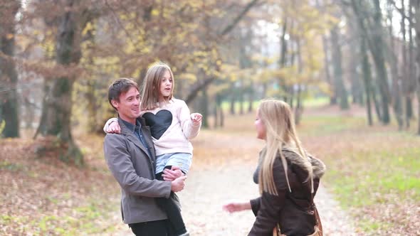 Family Walking In Park, Father Carries Daughter In His Arms And Mom Tickles Her