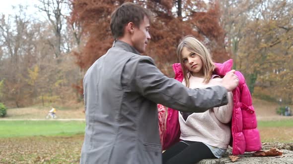 Father Helping Daughter Put On Jacket And Scarf In The Park