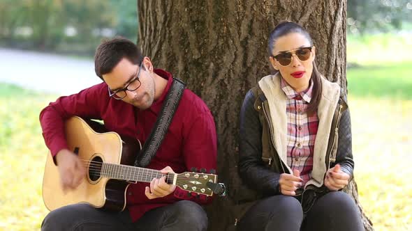 Woman Singing And Man Playing Guitar While Sitting On A Tree In Park 1