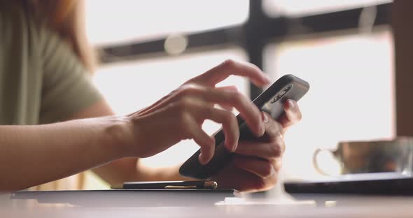 Closeup of a Girl with Smartphone in Her Hands Sitting at the Table Near the Window in Coffee Shop