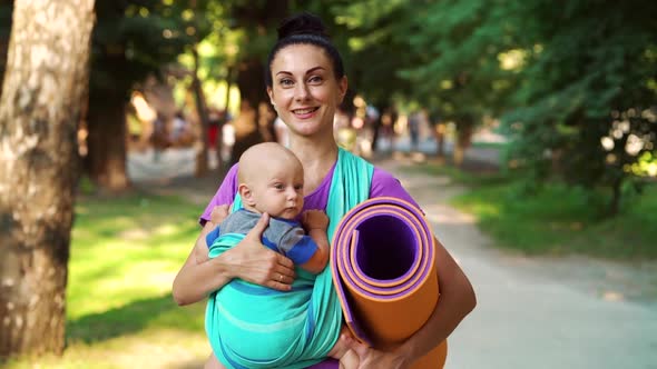 Happy mom with baby in sling going to yoga class in public park