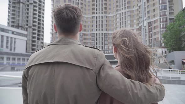 Brunette Young Woman Putting Head on Shoulder of Confident Man Hugging Her Outdoors. Back View of