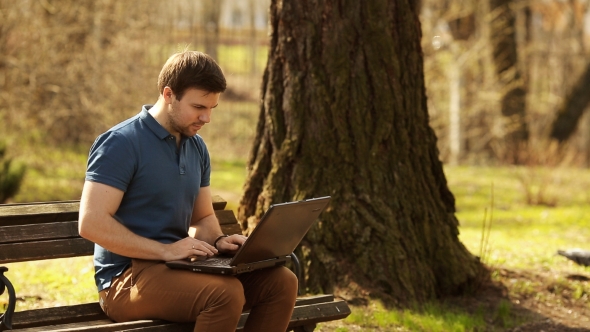Young Man Sitting On Park Bench Using Laptop
