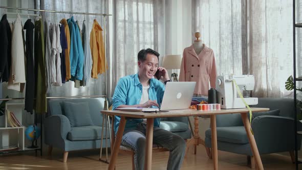 Asian Male Designer With Sewing Machine Talking On Smartphone While Designing Clothes On Laptop