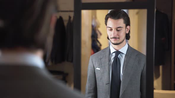 Satisfied young man trying on suit in front of mirror in fashion store.