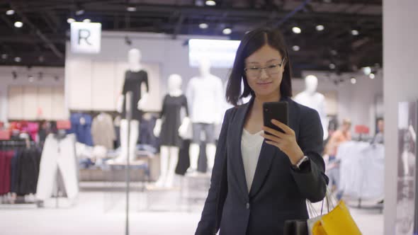 Delighted Asian Shopper Walking through Mall and Using Smartphone