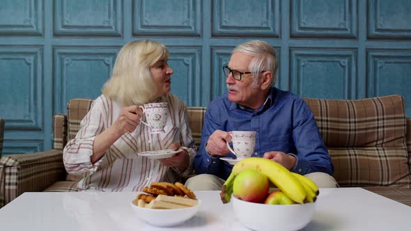 Senior Couple Holding Drinking Tea Talking Laughing Smiling Kissing Sitting on Couch at Home