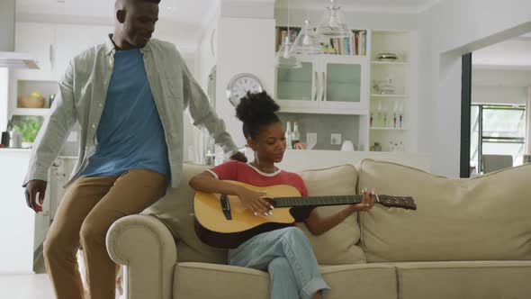 Video of happy african american couple sitting on sofa, singing and playing guitar