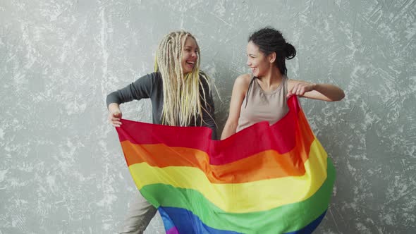 Positive Girlfriends Waving a LGBTQ Flag and Looking Into Each Other Eyes