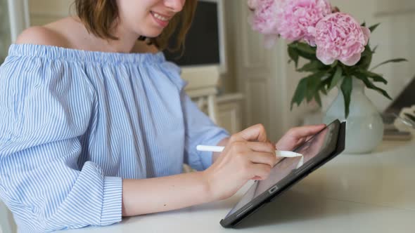 Young Cheerful Woman Working on the Tablet with Pencil in Modern Interior with Peonies on the