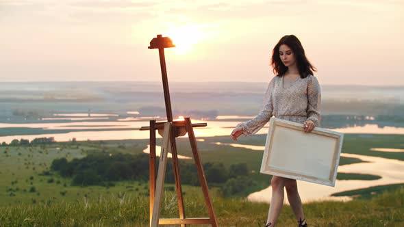 A Young Woman Artist in Light Dress Walks to an Easel Standing on the Sunset Field and Putting a