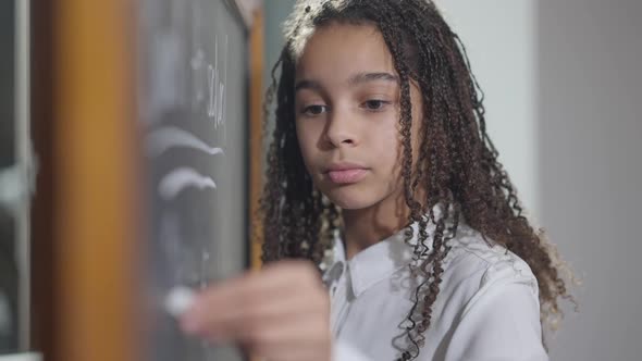 Serious Concentrated Lefthanded Schoolgirl Writing with Chalk on Blackboard in Classroom