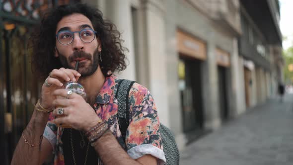 Smiling curly-haired bearded man in eyeglasses drinking juice and walking
