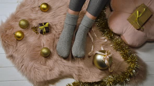 Woman in Gray Socks Sits on a Fur Skin Around Boxes with Gifts and Gold Colored Christmas