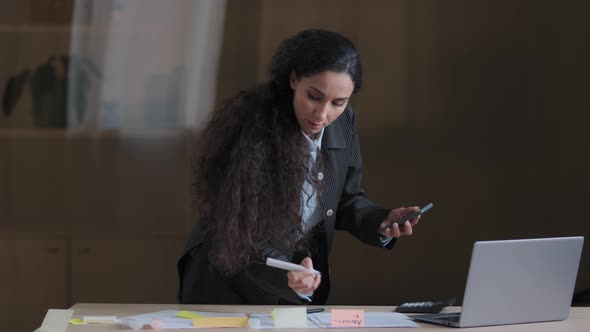 Focused Female Arabian Employee Designer Business Woman Standing at Office Desk Using Mobile Phone