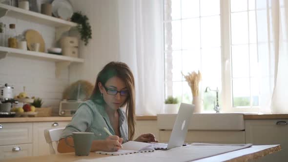Young Female Freelancer Ig Glasses Working on Laptop and Taking Notes in the Bright Kitchen