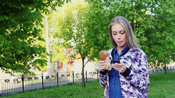 A Young Woman Walking Down The Street, Eating Fast Food Using a Mobile Phone