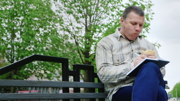 Man Eating Junk Food On a Park Bench, Writes Something In a Notebook