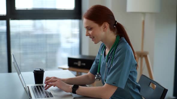 Cheerful Smiling Young Female Doctor in Blue Green Medical Uniform Typing on Laptop Computer Looking