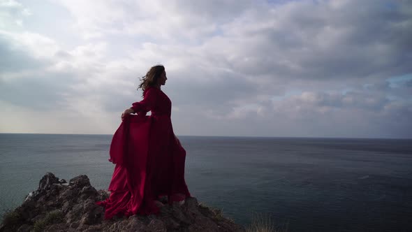 Side View a Young Beautiful Sensual Woman in a Red Long Dress Posing High Above the Sea on a