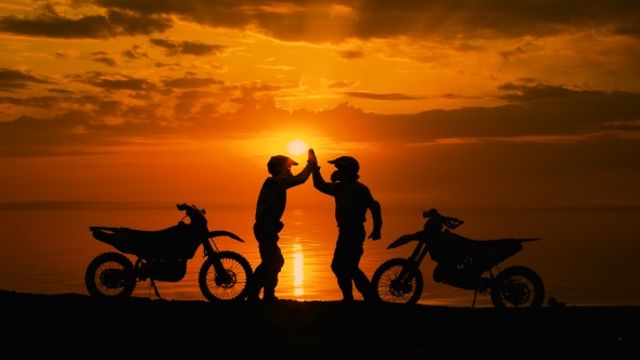 Two Motorcyclists At Sunset Communicate. They Are Naberegu River, Near His Motocross Bike. 