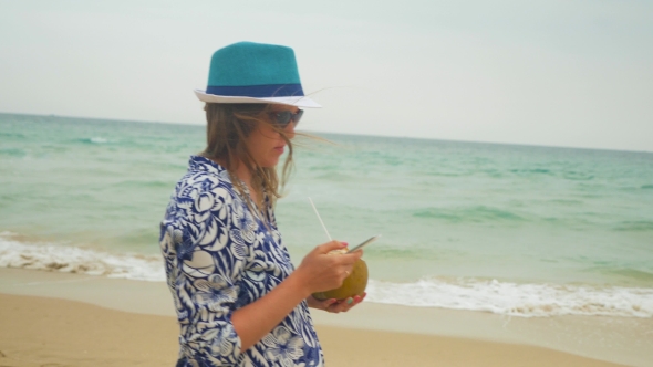 Pregnant Woman Walking Along The Sea With Coconut And Typing a Massage With a Cellphone
