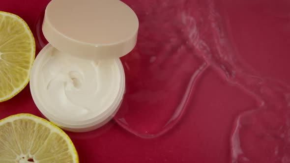 Closeup Cream Jar and Pieces of Fresh Lemons in Clean Water Splash Top View