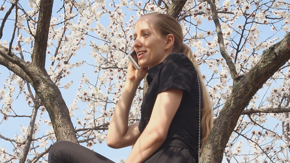 Girl Sitting on a Flowering Tree and Speaks by Phone