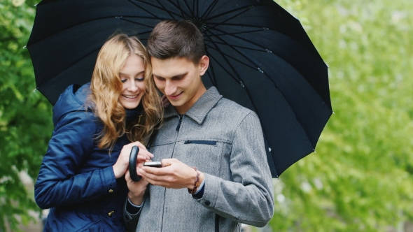 Young Couple Enjoys a Smartphone - Standing Under an Umbrella
