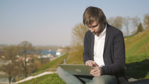 Portrait Of Young Man Using Laptop Outdoors Sitting On The Grass In Park