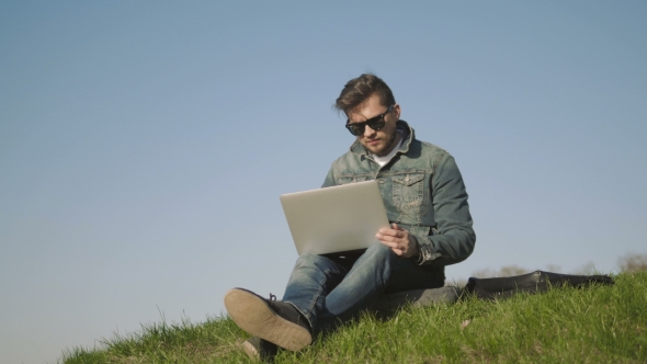 Young Man In Sunglasses Using Laptop Outdoors Sitting On The Grass In Park