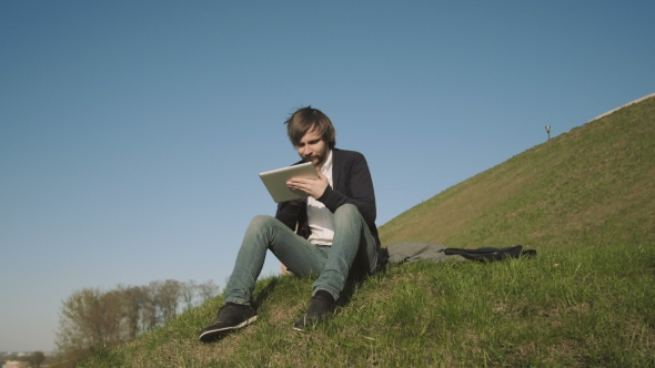 Handsome Hipster Using Tablet PC While Sitting On The Grass In The Park