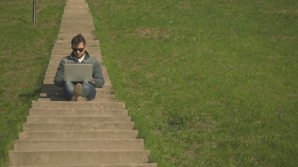 Young Hipster Man in Sunglasses Sitting on the Stairs Using Laptop