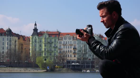 A Young Handsome Man Takes Photos with a Camera - a River and a Quaint Town in the Background