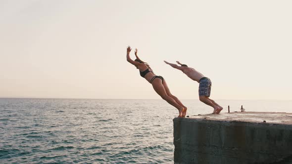 Young Woman and Man Synchronously Doing Frontflip From a Pier Into the Sea During Beautiful Sunrise