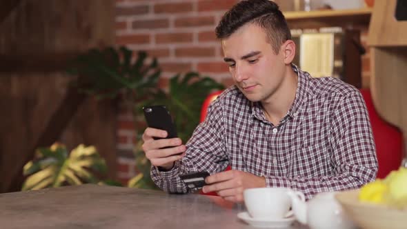 Handsome Man Making Purchase Online, Typing Credit Card Details on Black Smartphone in the Modern